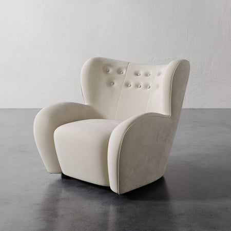 Lemieux et Cie Mondaine Lounge Chair in ivory with button-tufted back, sculptural upholstered armchair on dark concrete floor against white textured wall.