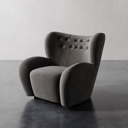 Lemieux et Cie Mondaine Lounge Chair in charcoal upholstery with button-tufted back and sculptural wingback form, on concrete floor against textured white walls.