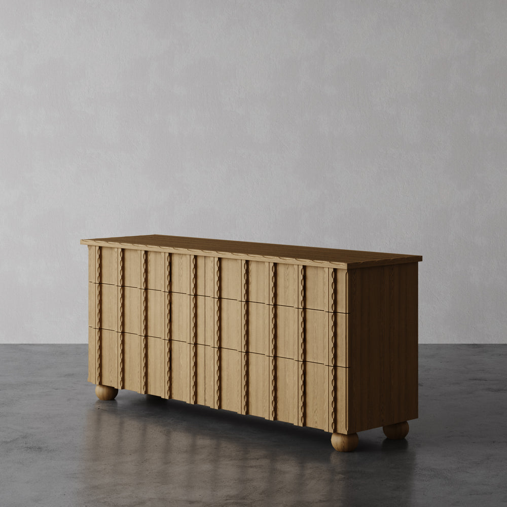 Lemieux et Cie Marchand 6-drawer dresser with scalloped carved relief drawer fronts and turned legs, shown on dark concrete flooring against neutral walls.
