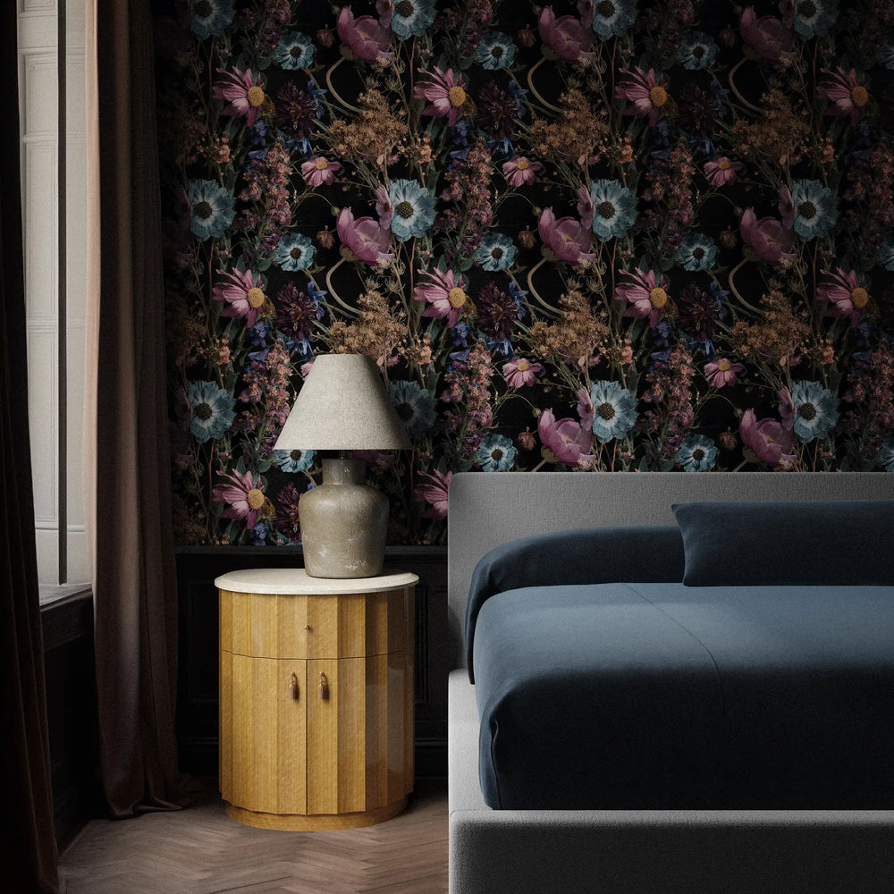Lemieux et Cie Midnight Meadow Botanical Wallpaper in violet, blue and rose on black ground, styled behind a side table and blue sofa in a bedroom with wood floors.