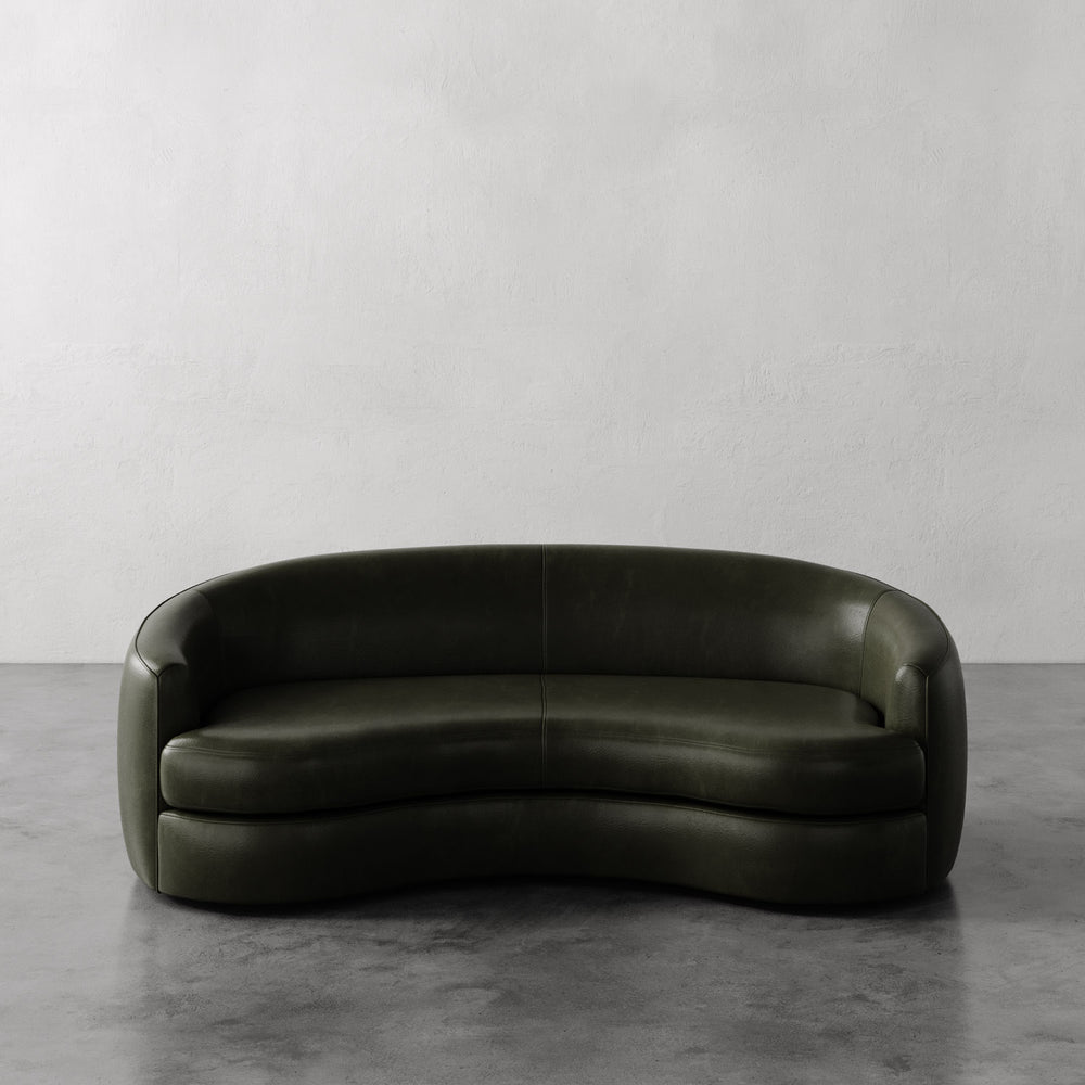 Lemieux et Cie Rulmann Sofa in deep olive leather, curved sculptural form with rounded arms, shown on gray concrete floor against white walls.