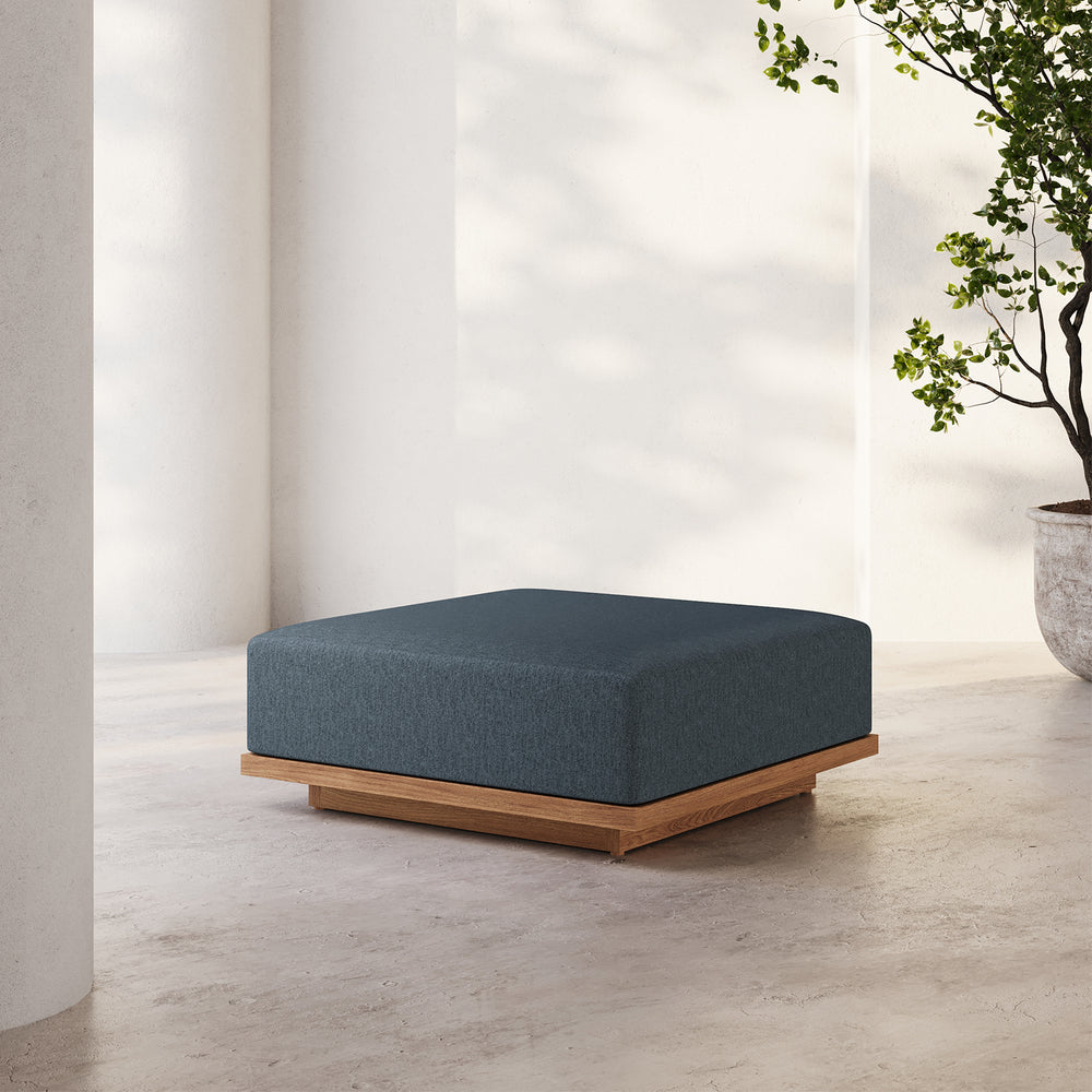 Lemieux et Cie S2 Teak Ottoman in deep blue upholstery with solid teak base, outdoor ottoman on concrete floor in sunlit white-walled space.