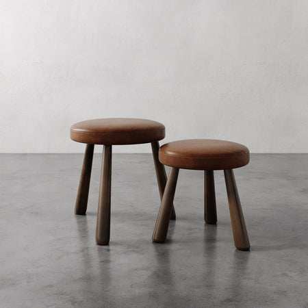 Lemieux et Cie Madeleine Leather Stool in tobacco leather with tapered legs, low stools shown on polished concrete floor against textured plaster walls.