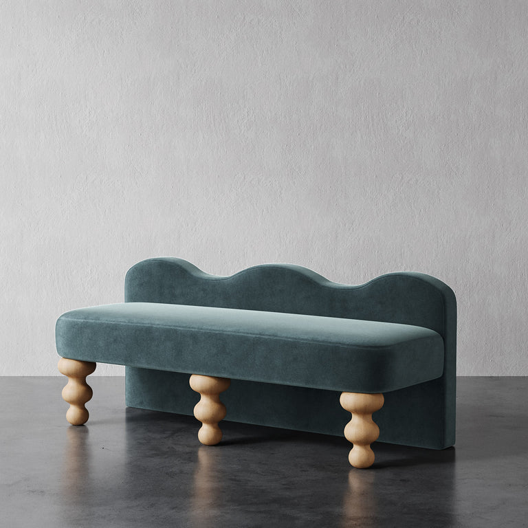Lemieux et Cie Celine Custom Upholstered Bench in deep teal velvet with hand-turned legs, scalloped back bench on dark stone floor against textured plaster wall.