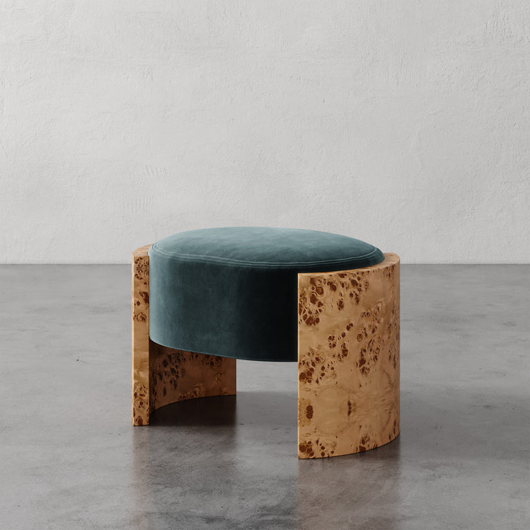 Lemieux et Cie Benoit Ottoman with deep teal upholstered seat and curved oak frame, low-profile stool shown on concrete floor against white textured wall.