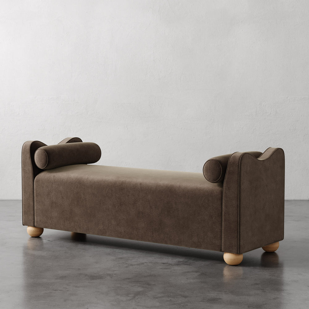 Lemieux et Cie Danielle Upholstered Bench in warm taupe with cylindrical bolster arms and rounded feet, shown on dark concrete floor against white plaster wall.