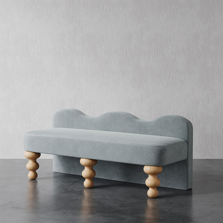Lemieux et Cie Celine Custom Upholstered Bench in pale blue with scalloped back and hand-turned wood legs, shown on dark stone floor against textured white walls.