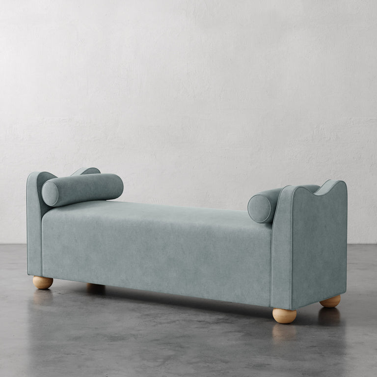 Lemieux et Cie Danielle Upholstered Bench in soft blue-green, low bench with cylindrical bolster arms and rounded legs, shown on polished concrete floor against neutral plaster walls.