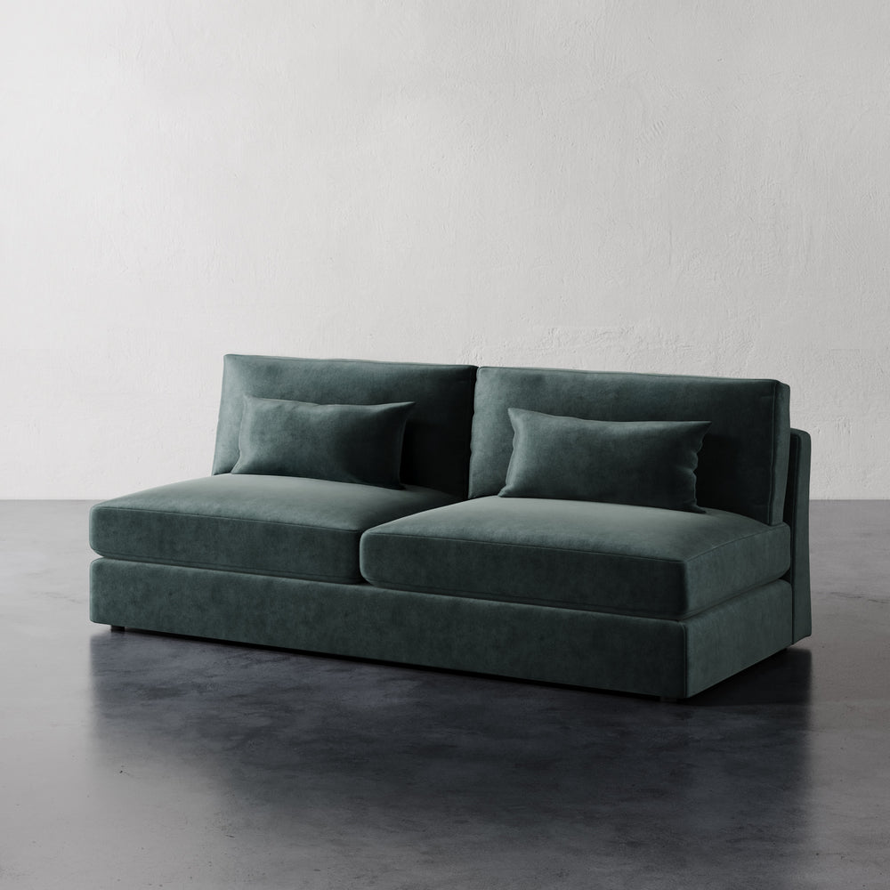 Lemieux et Cie Lacroix Armless Sofa in deep green velvet, modular sofa with rectilinear form and bolster pillows, shown on dark concrete floor against textured white walls.