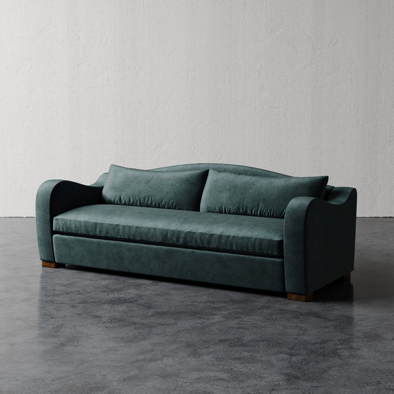 Lemieux et Cie Elodie Sofa in deep teal, bench seat sofa with rounded arms and plush cushions, shown on concrete floor against textured white plaster wall.