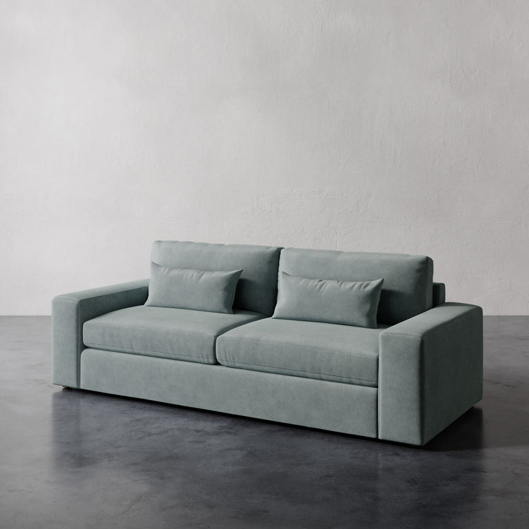Lemieux et Cie Lacroix Sofa in sage green, two-cushion sofa with bolster pillows, shown on dark polished concrete floor against textured plaster walls.