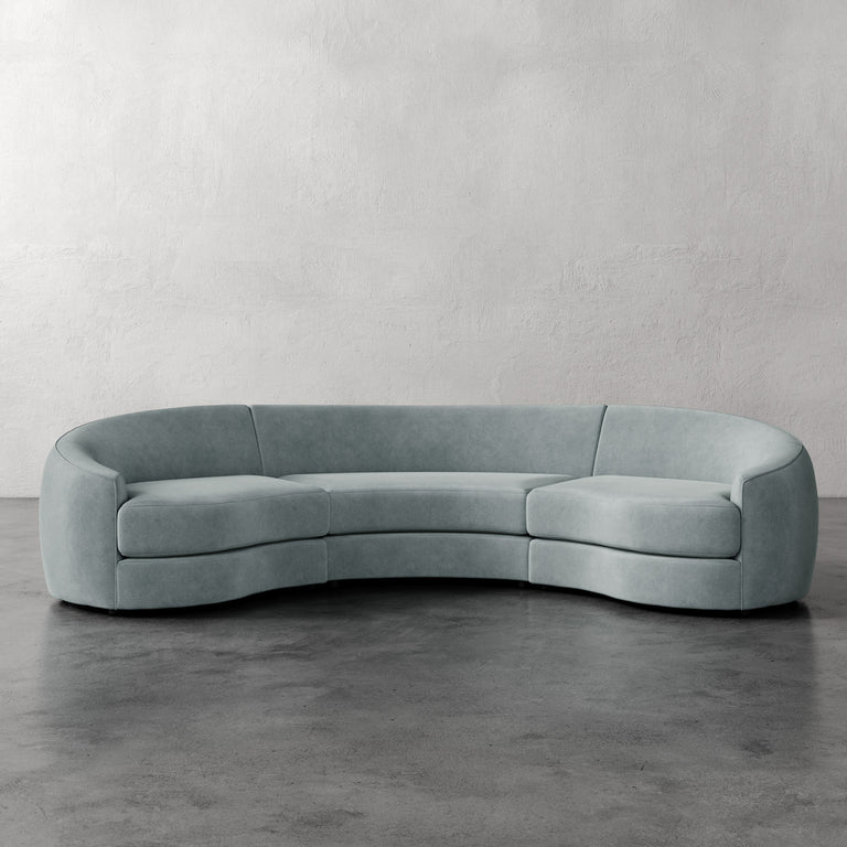 Lemieux et Cie Rulmann 3-Piece Sectional in soft sage, curved three-piece sofa with rounded arms, shown on concrete floor against textured plaster wall.