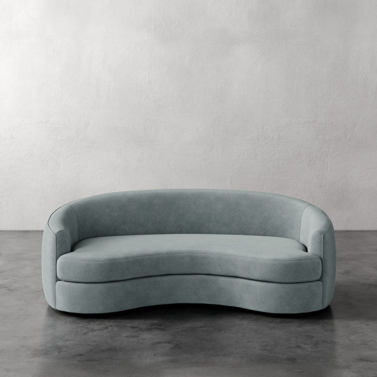 Lemieux et Cie Rulmann Sofa in soft sage green, curved organic sofa with rounded back, shown on dark concrete floors against textured white plaster walls.