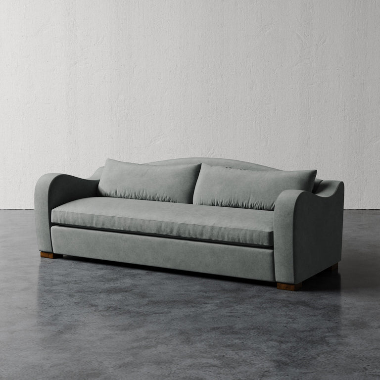 Lemieux et Cie Elodie Sofa with bench seat in soft gray, low-profile sofa with rounded arms and two cushions, shown on concrete floor against textured plaster walls.