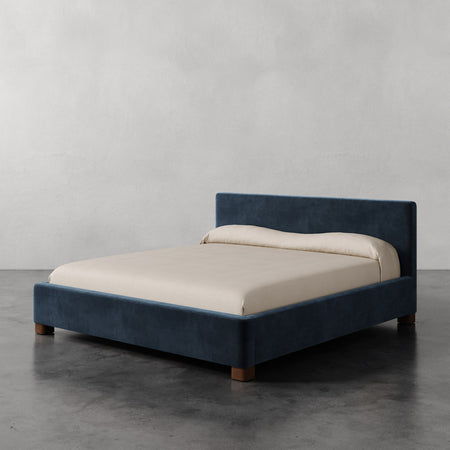 Lemieux et Cie Gérard Bed in deep blue upholstery with ivory bedding, low-profile platform bed on dark concrete floor against neutral plaster walls.