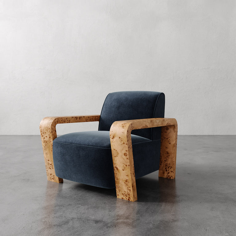 Lemieux et Cie Dumas Chair in dark blue upholstery with blonde burl curved arms, lounge chair shown on polished concrete floor against white walls.
