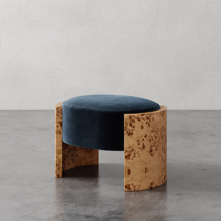 Lemieux et Cie Benoit Ottoman in deep blue velvet with natural burl wood frame, low ottoman on polished concrete floor against textured plaster walls.