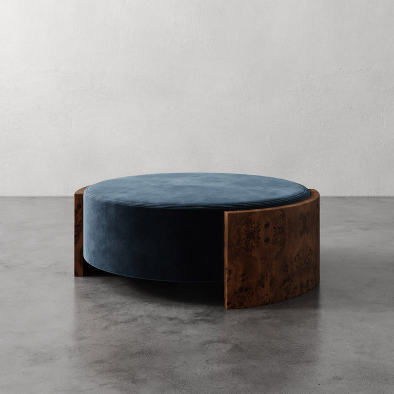 Lemieux et Cie Benoit Coffee Table Ottoman with cylindrical blue velvet cushion and curved wire brushed oak frame on concrete floor against neutral plaster walls.