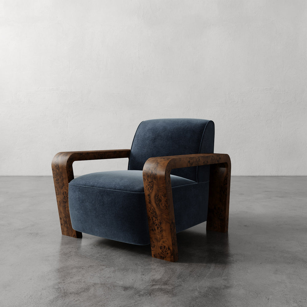 Lemieux et Cie Dumas Chair in dark blue velvet with curved burl wood arms, lounge chair shown on concrete floor against textured plaster wall.