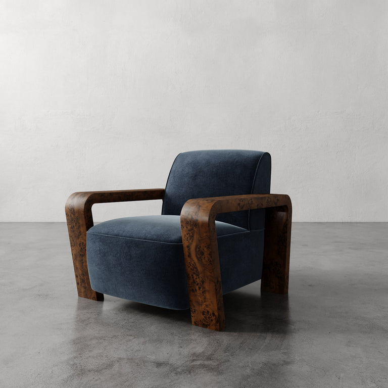 Lemieux et Cie Dumas Chair in dark blue velvet with curved burl wood arms, lounge chair shown on concrete floor against textured plaster wall.