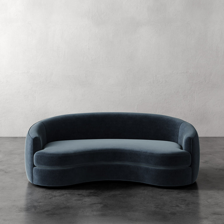 Lemieux et Cie Rulmann Sofa in deep blue, curved organic-form sofa with rounded back and arms, on dark concrete floor against textured plaster walls.