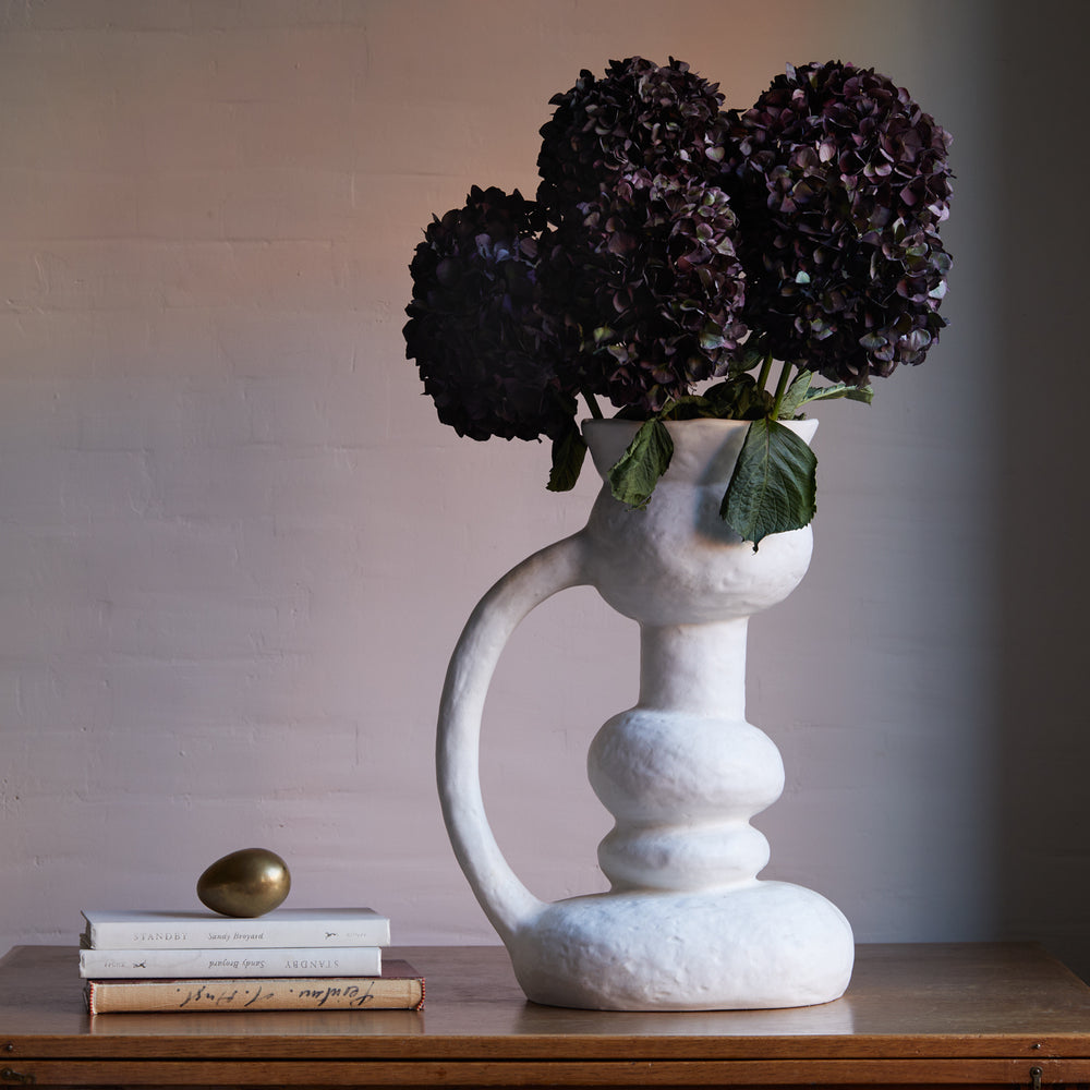 Lemieux et Cie Remi Vase in white, sculptural ceramic vessel with organic curved form, holding dark hydrangeas on a wood surface with stacked books.