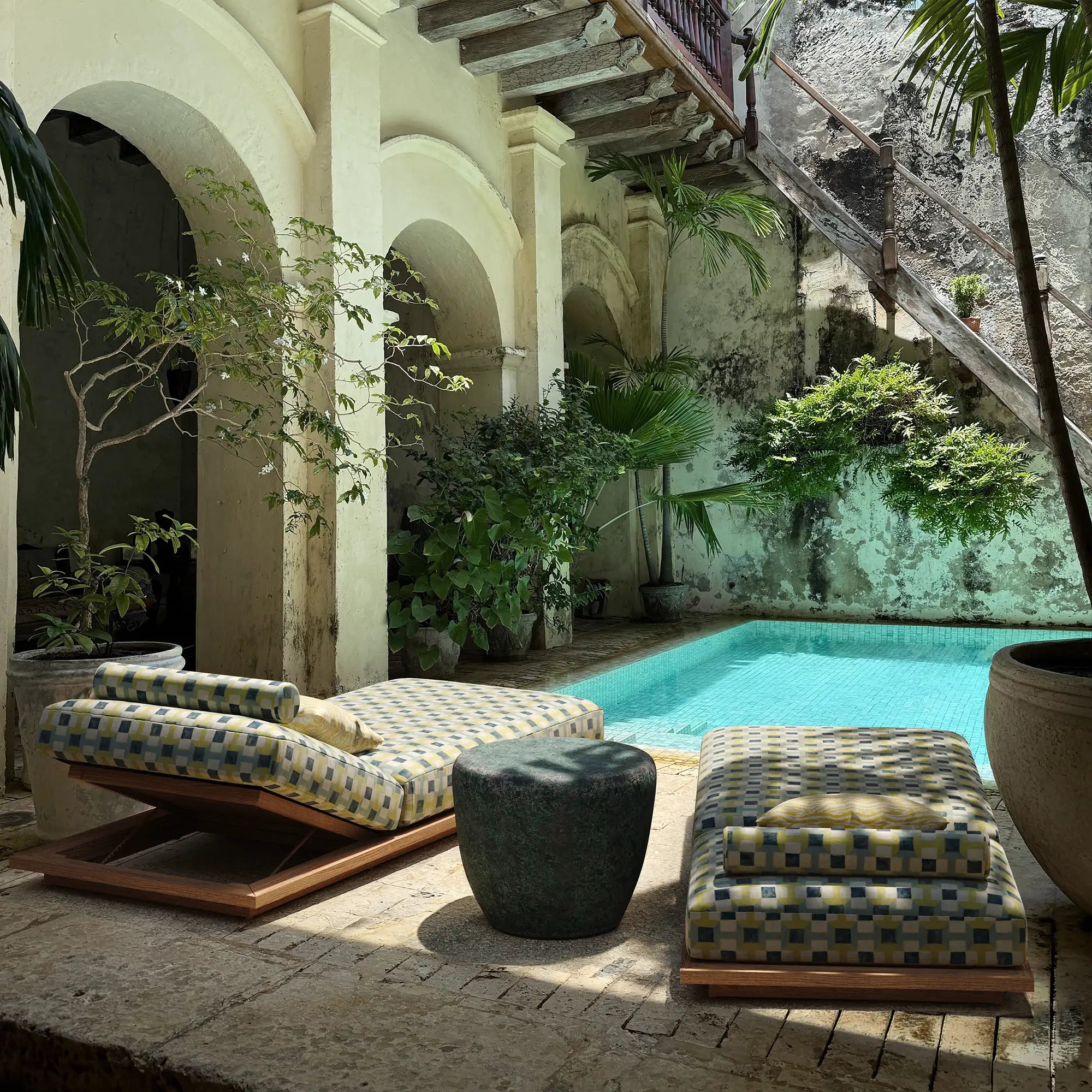 Outdoor patio with lounge chairs, pool, and plants in a sunlit courtyard. With the Taiko Stool / End Table by Lemieux et Cie in Oxidized Jade reactive glaze