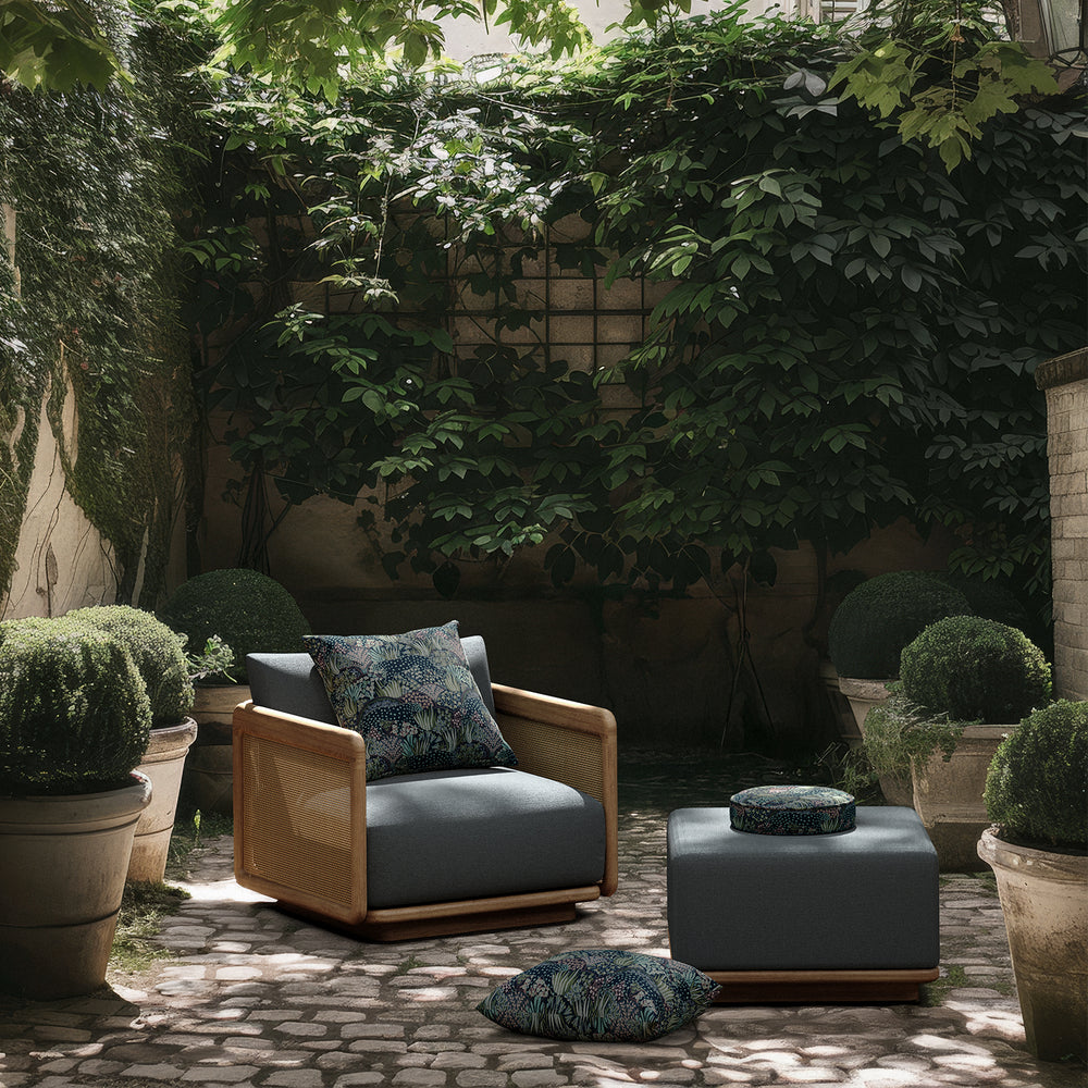 Lemieux et Cie Dock Teak Ottoman in charcoal upholstery with teak base, outdoor ottoman styled on stone patio with lush greenery and botanical pillows.