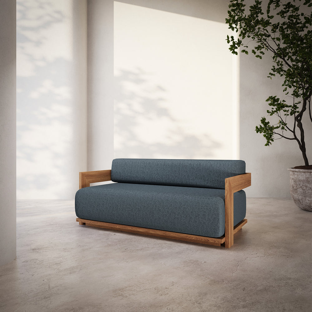 Lemieux et Cie S2 Teak 62" Sofa in slate blue upholstery with warm teak frame, shown on concrete floor against white walls in soft daylight.