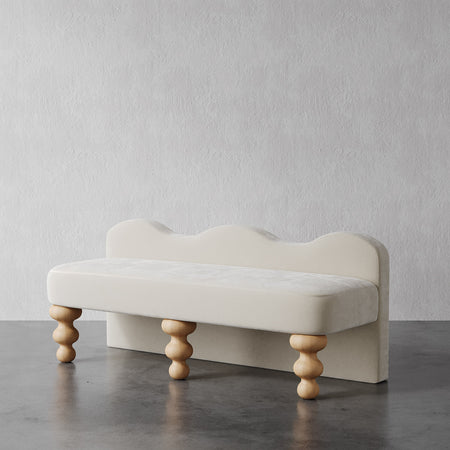 Lemieux et Cie Celine Custom Upholstered Bench in ivory with scalloped back and hand-turned wooden legs, shown on dark concrete floor against textured plaster wall.