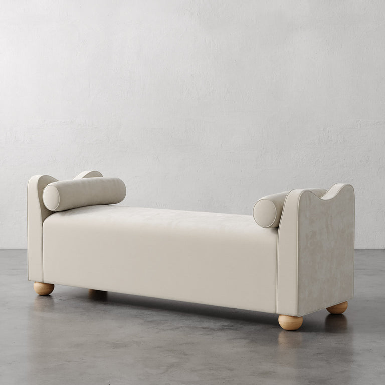 Lemieux et Cie Danielle Upholstered Bench in ivory with sculptural armrests and bolster pillows, rounded feet, shown on concrete floor against neutral plaster walls.