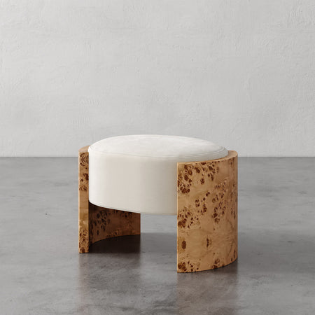 Lemieux et Cie Benoit Ottoman in cream with burled wood curved base, low-profile ottoman on gray concrete floor against white textured wall.