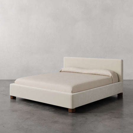Lemieux et Cie Gérard Bed, low-profile platform bed with wrapped headboard and rounded block feet, shown on polished concrete floor against neutral plaster walls.