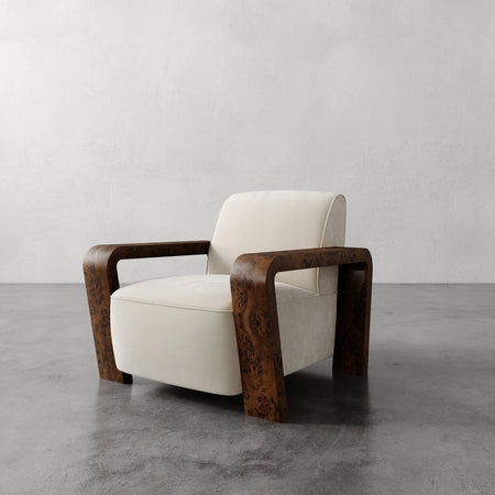 Lemieux et Cie Dumas Chair with walnut burl arms and ivory upholstery, lounge chair shown on gray concrete floor against textured plaster walls.