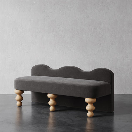 Lemieux et Cie Celine Custom Upholstered Bench in charcoal with scalloped back and hand-turned legs, shown on dark stone surface against textured plaster wall.