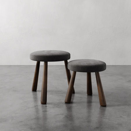 Lemieux et Cie Madeleine Stool with charcoal upholstered seat and dark tapered legs, shown as nesting pair on concrete floor against white walls.