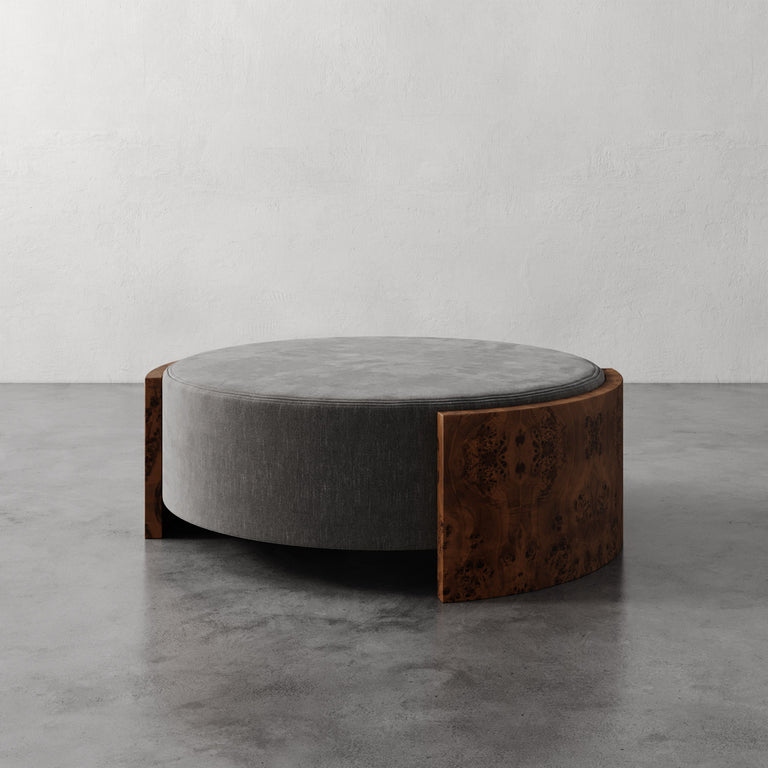 Lemieux et Cie Benoit Coffee Table Ottoman in gray upholstery with wire brushed oak curved frame, low-profile ottoman on concrete floor against white plaster walls.