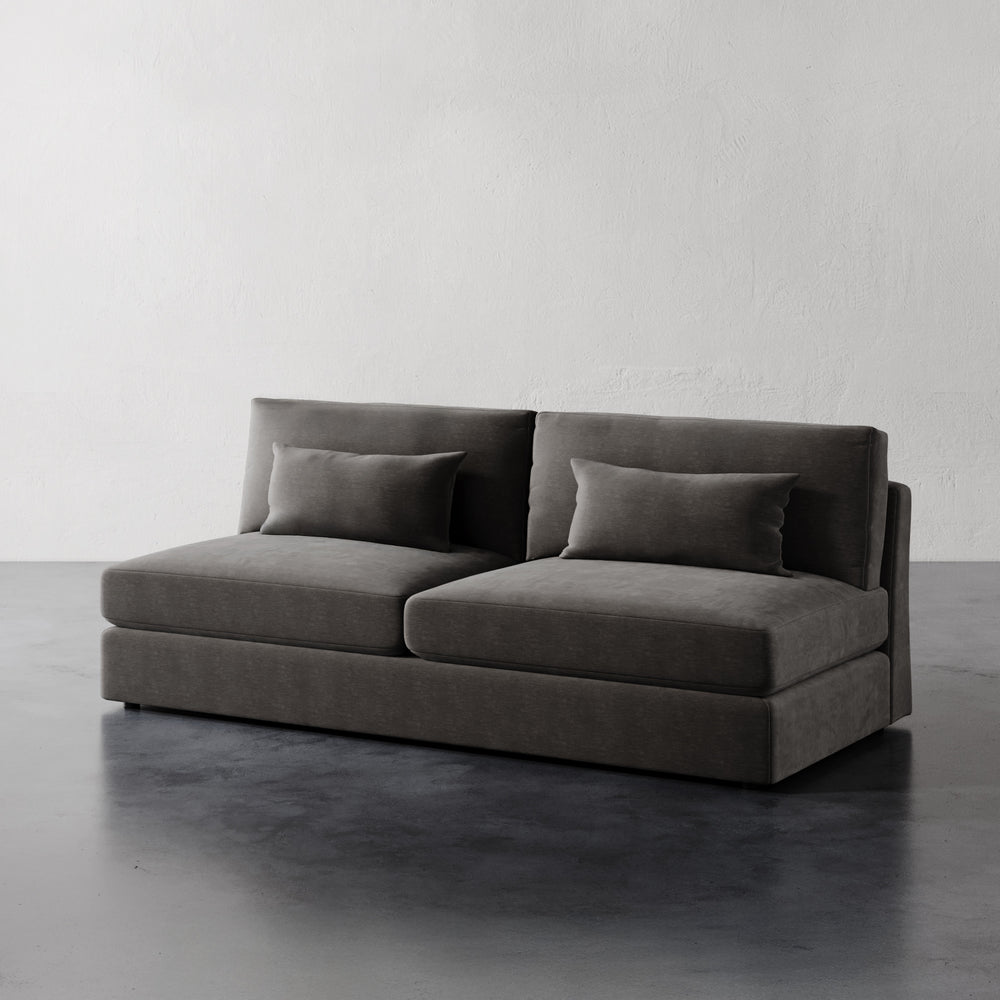 Lemieux et Cie Lacroix Armless Sofa in charcoal upholstery, modular sofa with bolster pillows, shown on dark concrete floor against white plaster walls.