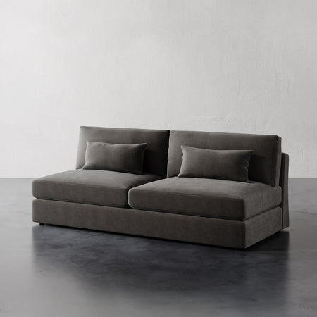 Lemieux et Cie Lacroix Armless Sofa in charcoal upholstery, modular sofa with bolster pillows, shown on dark concrete floor against white plaster walls.