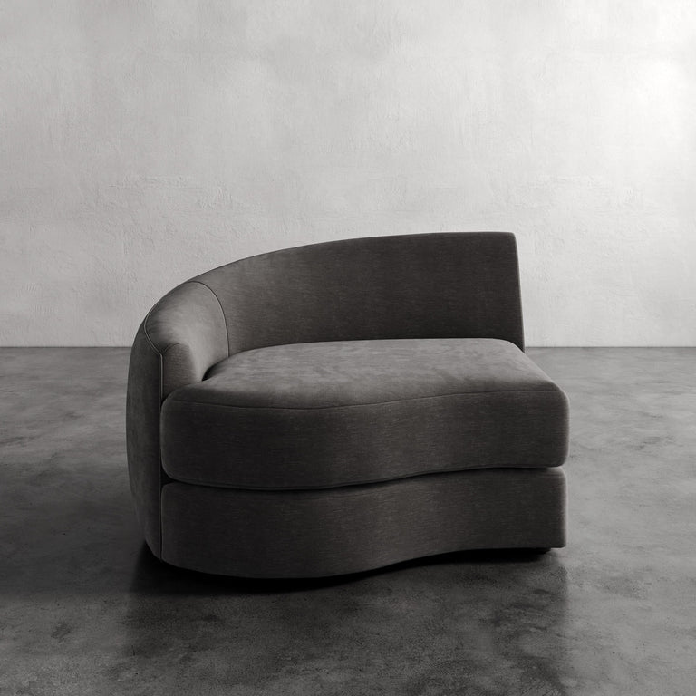 Lemieux Et Cie Rulmann Left Arm Chair Gray curved sofa in a minimalistic room with concrete floor and wall.