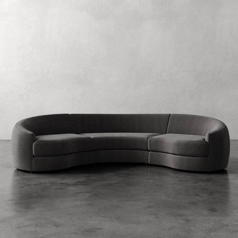Lemieux et Cie Rulmann 3-Piece Sectional, curved sectional sofa in charcoal with rounded arms and low profile, on polished concrete floor against textured plaster walls.