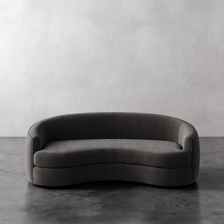 Lemieux et Cie Rulmann Sofa, curved sculptural form in charcoal gray, shown on dark concrete floor against textured plaster walls.