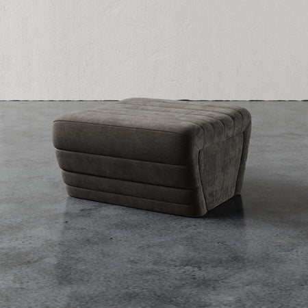 Lemieux et Cie Brunoy Ottoman in charcoal with channel-tufted upholstery, low-profile ottoman on stone floor against textured plaster wall.