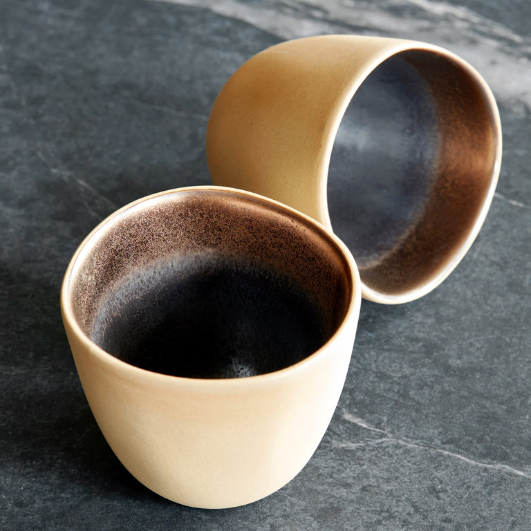 Two stoneware mugs with a reactive glaze in brown and black colors, one with the interior facing up and the other with the exterior showing a raw tan clay texture.