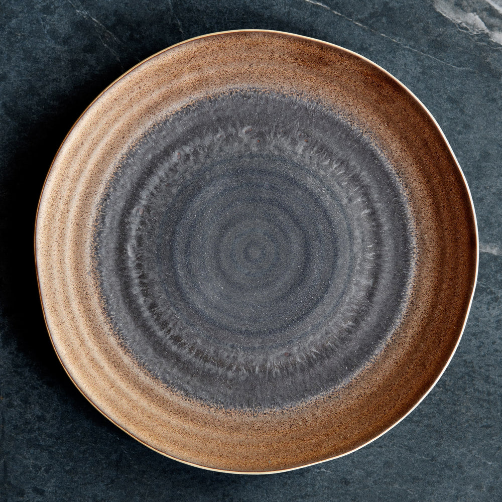 Lemieux et Cie Akita Stoneware Dinner Plate with reactive glaze in copper and charcoal concentric pattern, shown on dark stone surface.