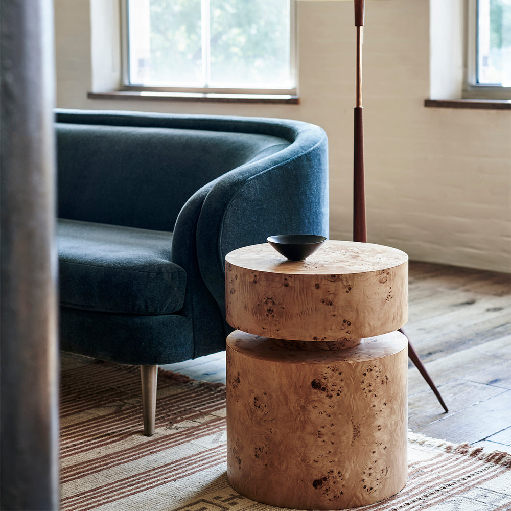 Lemieux et Cie Avenir Side Table in burl, cylindrical stacked side table with dark bowl, beside blue velvet chair on striped rug in soft daylit room.