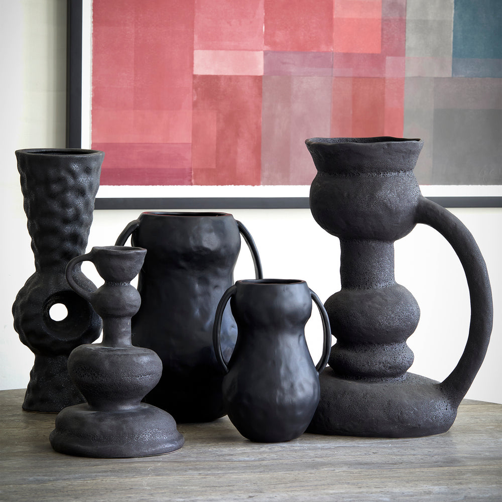 Lemieux et Cie Victoire Vase in black, sculptural ceramic vessels with organic forms and textured handles, grouped on dark wood surface with blush abstract art beyond.
