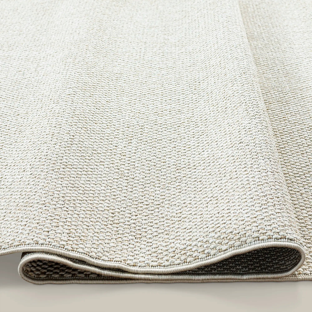 Lemieux et Cie Provence Outdoor Rug in cream melange, woven rug shown rolled to reveal textured surface and bound edge on neutral backdrop.