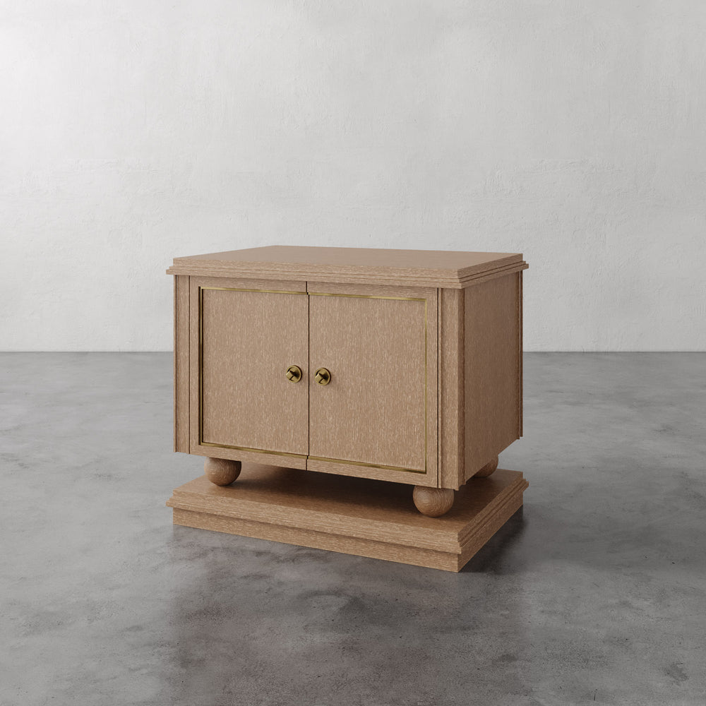 Lemieux et Cie Corvaux Nightstand in cerused oak with brass hardware and inlay, sculptural nightstand with turned feet shown on concrete floor.