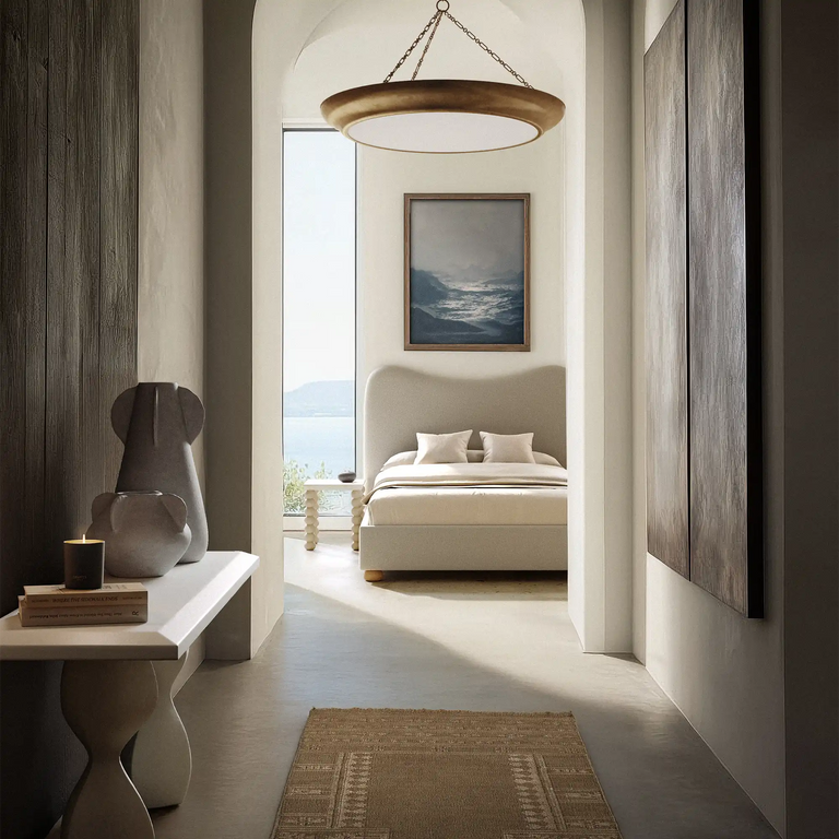 Lemieux et Cie Lenoir Chandelier in matte finish, sculptural iron bowl form suspended above an upholstered bed in a light-filled bedroom with neutral walls and stone flooring.